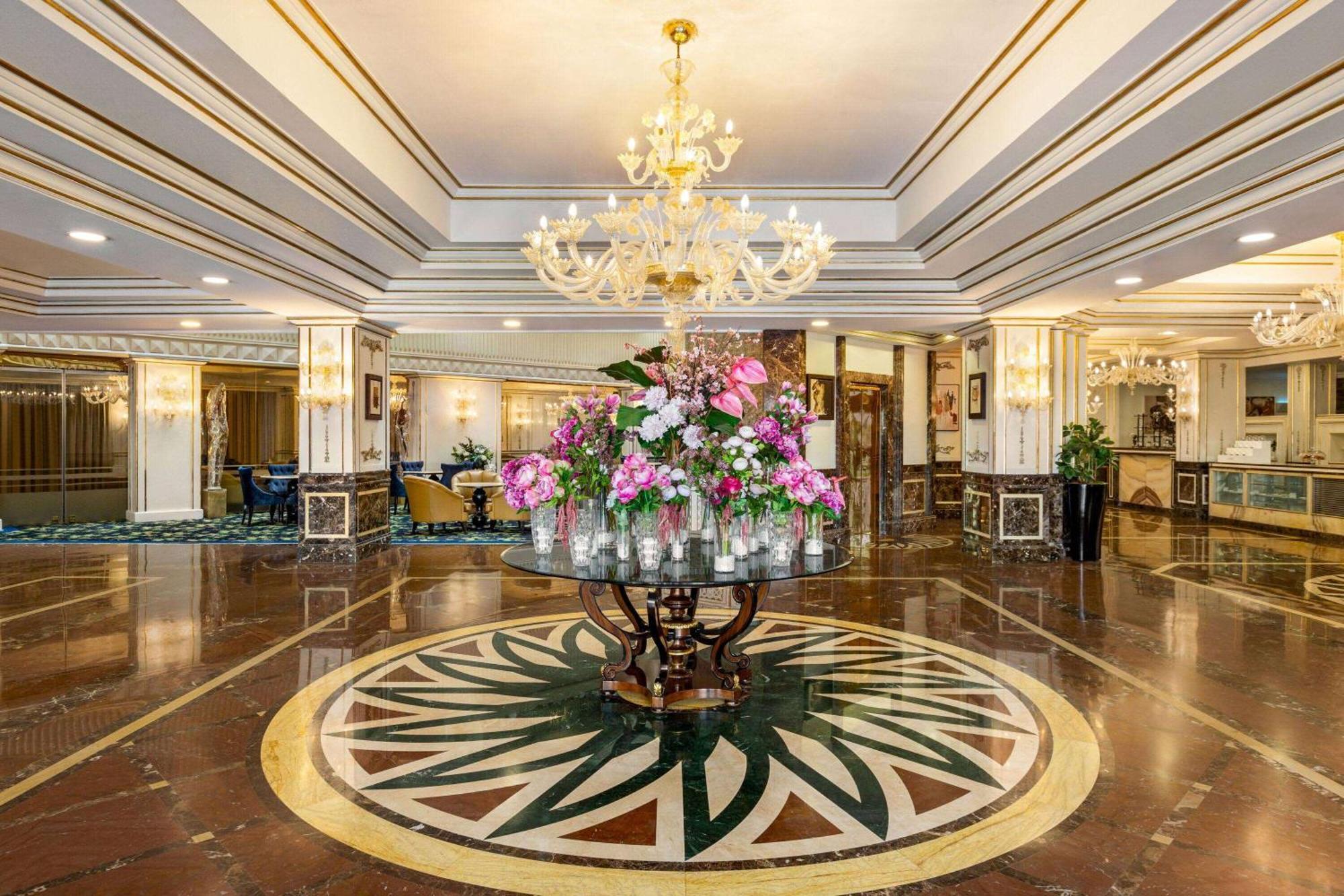 Grand Regency Doha, Trademark Collection By Wyndham Exterior photo The photo shows a luxurious hotel lobby featuring an elegant chandelier hanging from the ceiling. In the center, there is a large round table adorned with an arrangement of vibrant flowers. The floor showcases intricate marble patterns, and the overa