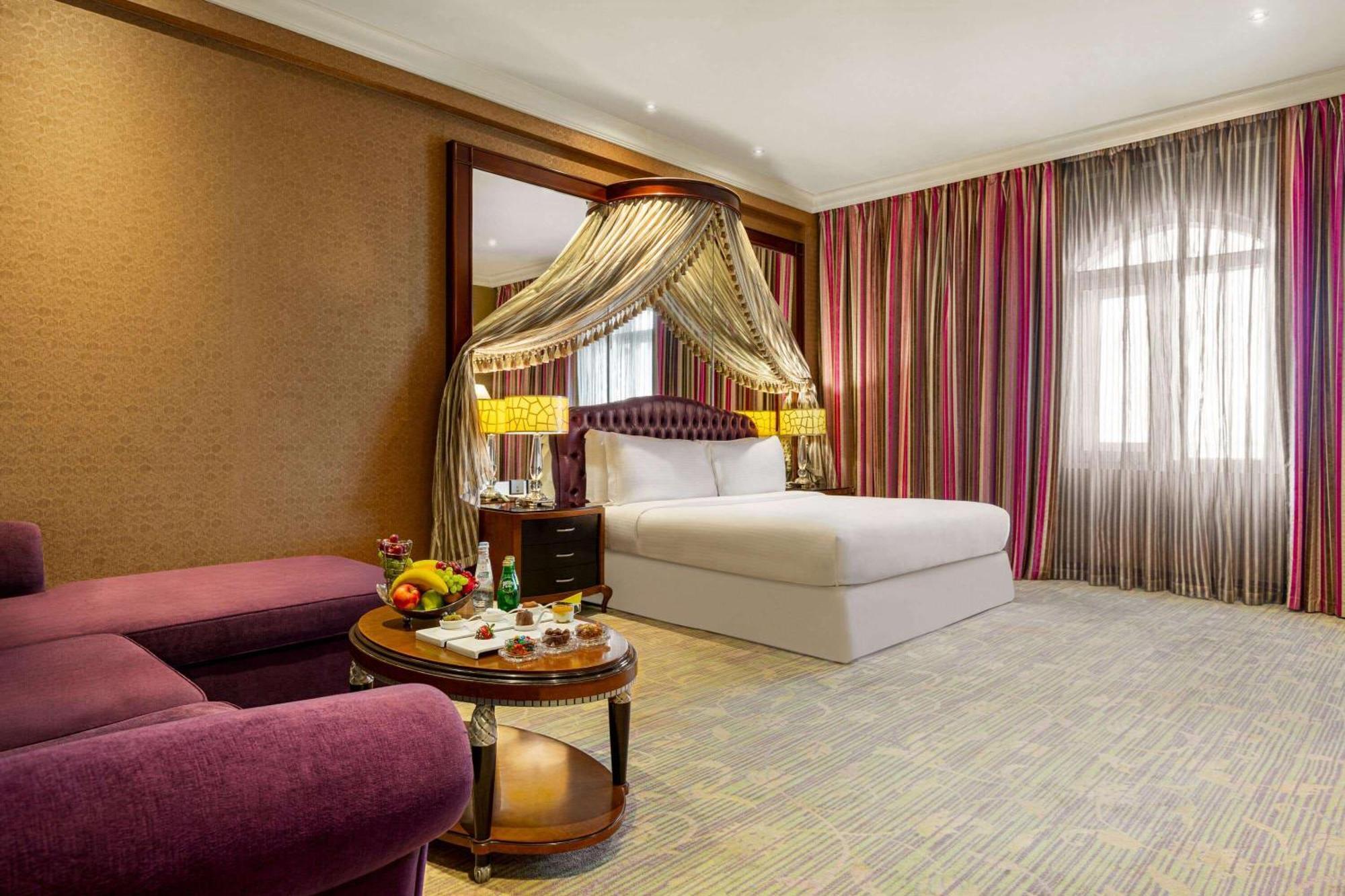 Grand Regency Doha, Trademark Collection By Wyndham Exterior photo The photo shows a luxurious hotel room featuring a king-sized bed with a decorative canopy. The room is elegantly designed with a mix of rich colors and textures. To the left, there's a plush purple seating area with a small round table that displays