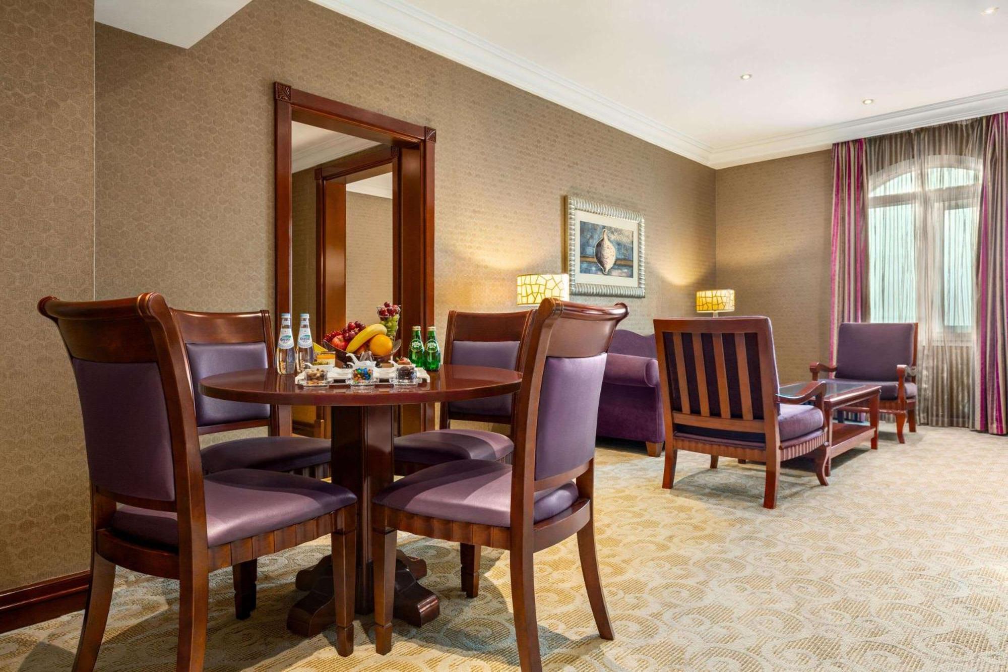 Grand Regency Doha, Trademark Collection By Wyndham Exterior photo The photo depicts a well-furnished hotel room or lounge area. In the foreground, there is a round table surrounded by four purple upholstered chairs, with a selection of fruits and beverages on the table. To the right, there are two wooden armchairs,